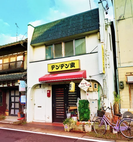 A narrow white shop has a red awning and yellow sign above potted plants and a purple bicycle.