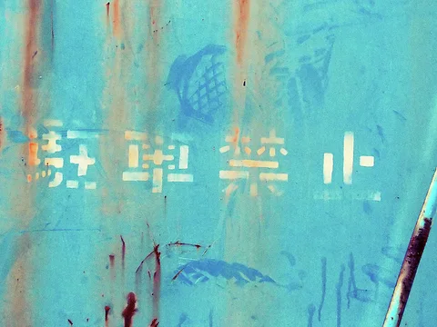 Faded pale Japanese characters and blue illustrations show through rust streaks on a turquoise metal panel.