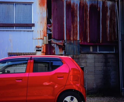 A bright red compact car is parked against patched corrugated walls streaked with rust.