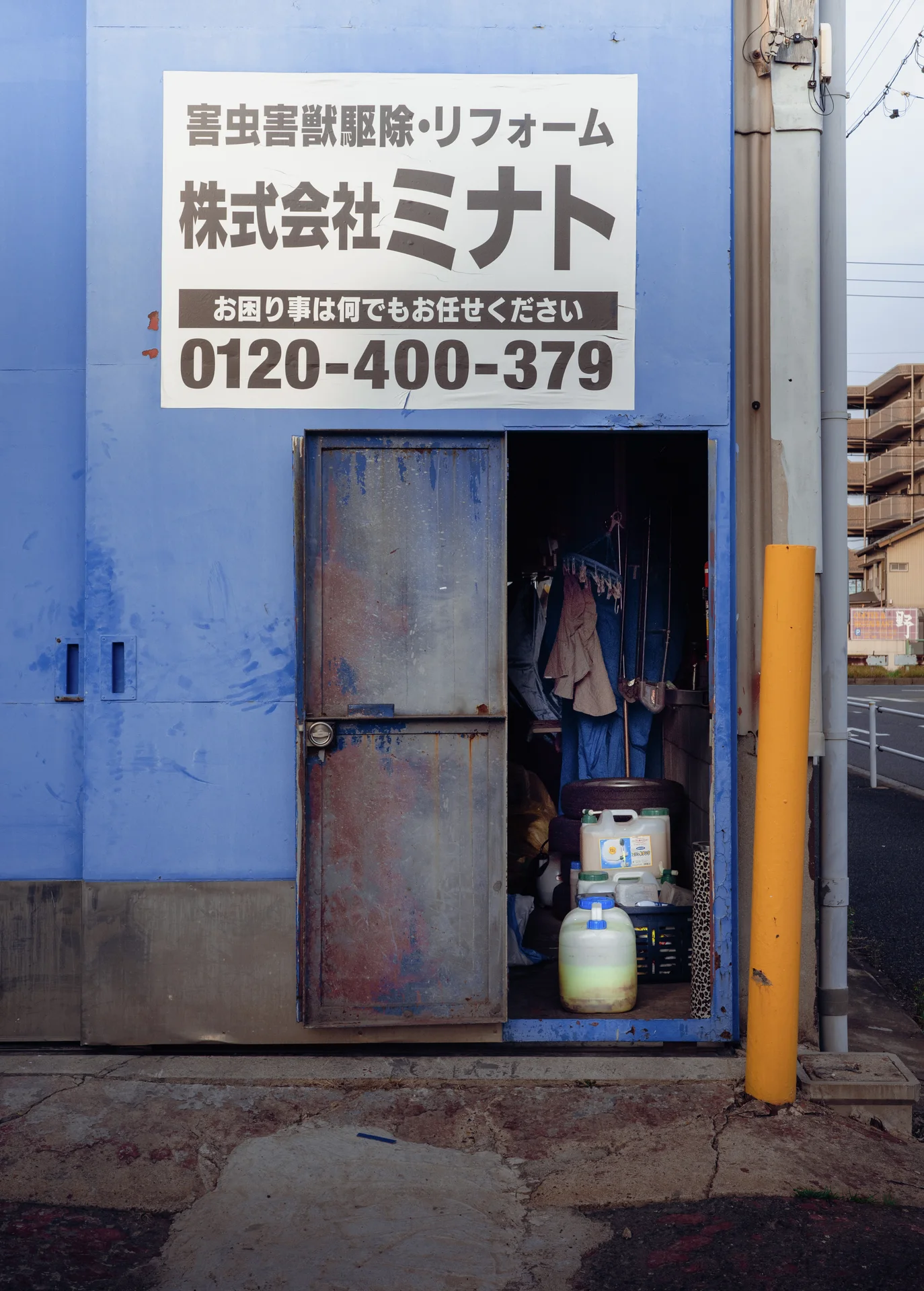 A small open door in a blue wall reveals stored containers beneath a white Japanese business sign.