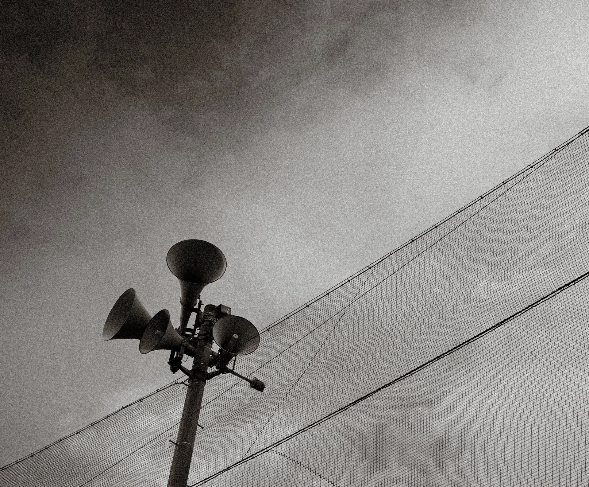 Horn loudspeakers rise beside overhead wires and tall netting against a monochrome sky.