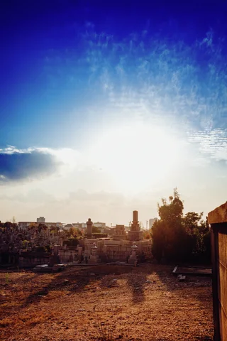 Bright low sun shines above an open dirt lot bordered by trees and distant buildings beneath blue sky.