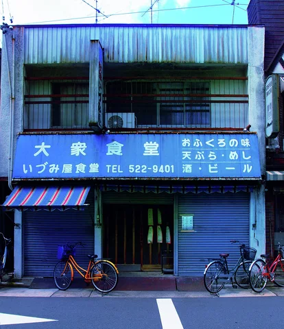 A blue restaurant facade has a broad sign, striped awning, closed shutters, and parked bicycles.