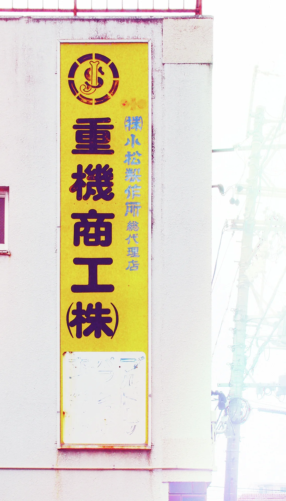 A narrow yellow vertical sign with dark characters hangs against a pale exterior wall.