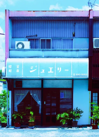 A turquoise storefront has pale Japanese lettering, curtained doors, potted plants, and a narrow upper balcony.