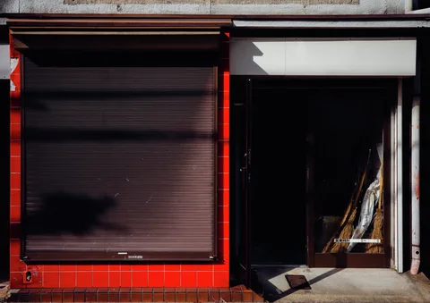 A dark closed shutter framed by red tiles stands beside a shadowed doorway containing leaning boards.