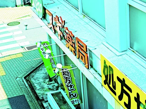 Raised orange pharmacy lettering and green banners project above a turquoise storefront beside a marked intersection.