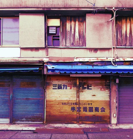 Weathered wooden upper windows overlook colorful closed storefront shutters beneath a blue awning.