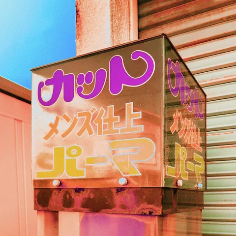 Purple and orange Japanese lettering covers a reflective projecting box sign beside a closed metal shutter.