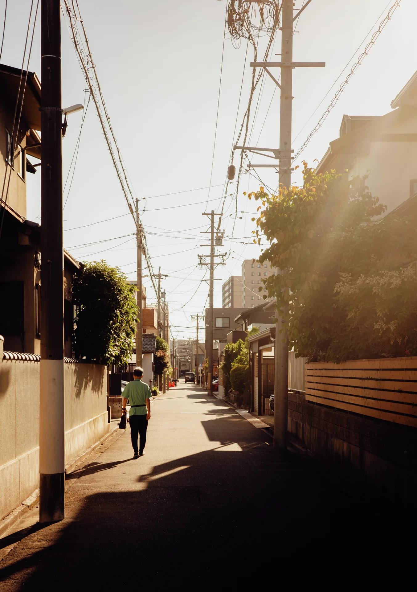 A pedestrian walks along a narrow sunlit residential lane beneath rows of overhead cables.
