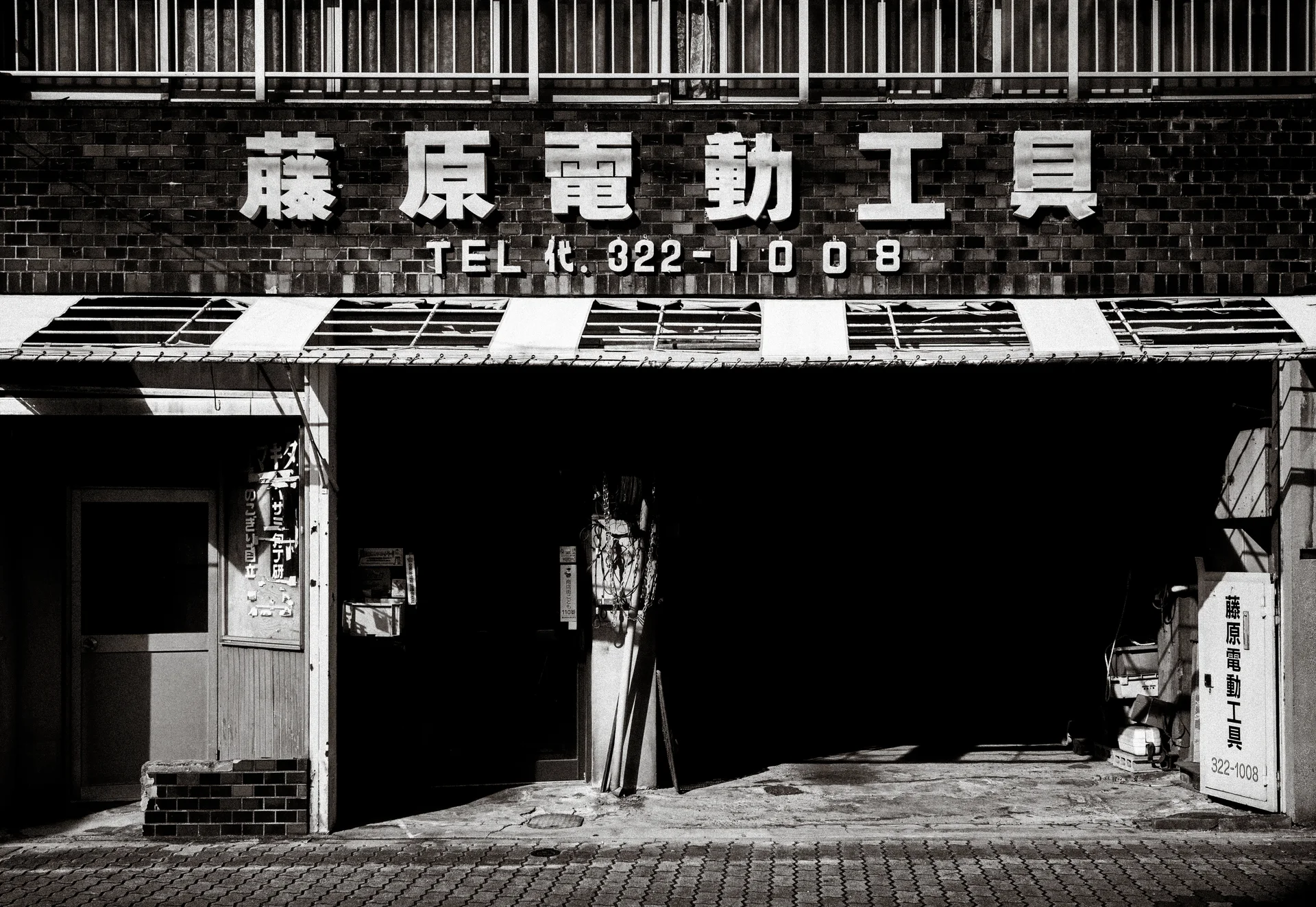 Raised white lettering spans a brick workshop facade above a deeply shadowed opening in monochrome.