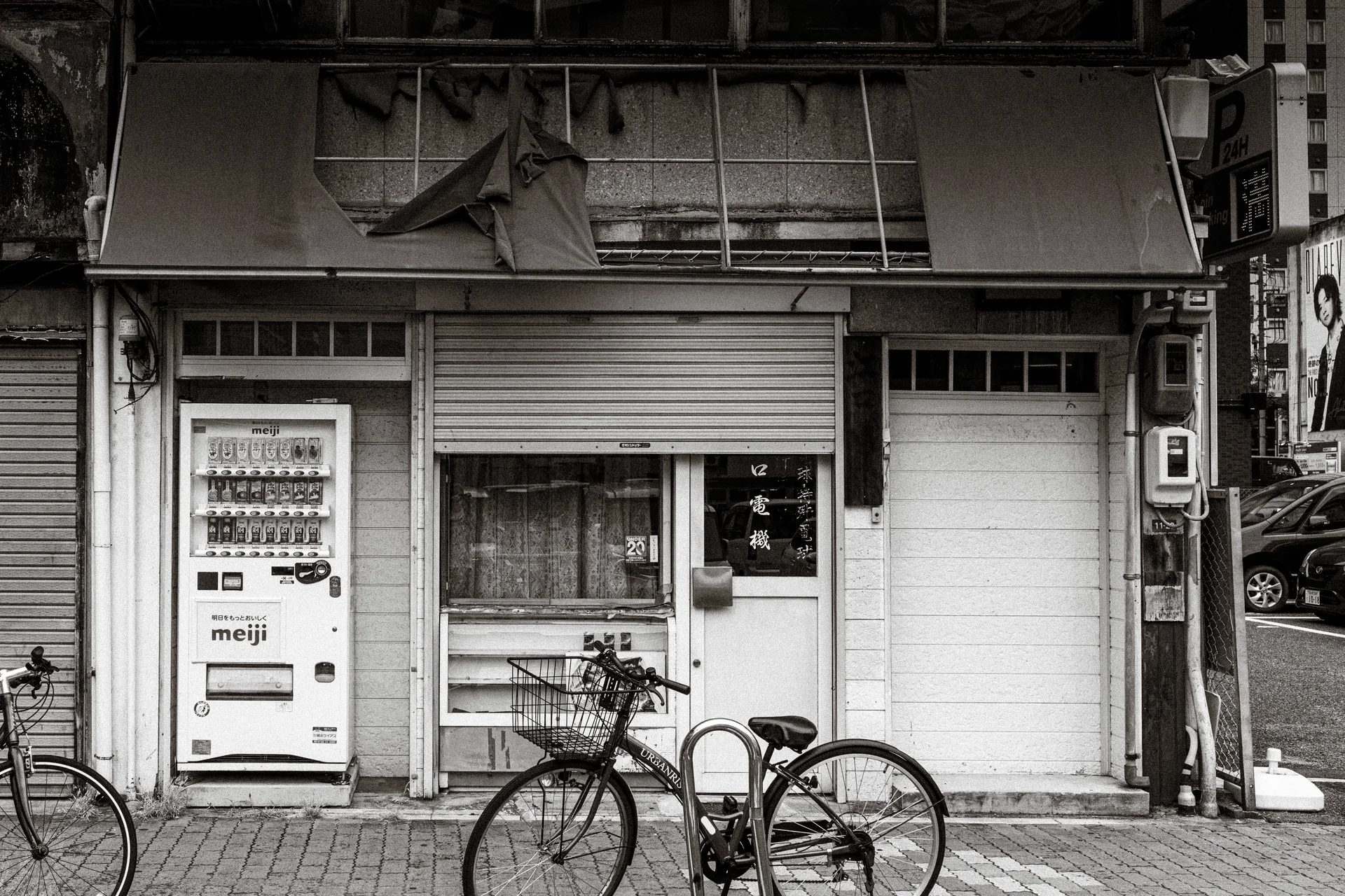 Bicycles stand outside an aging shuttered storefront beside a vending machine in black and white.