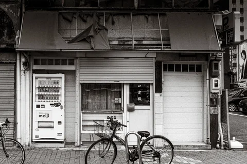 Bicycles stand outside an aging shuttered storefront beside a vending machine in black and white.