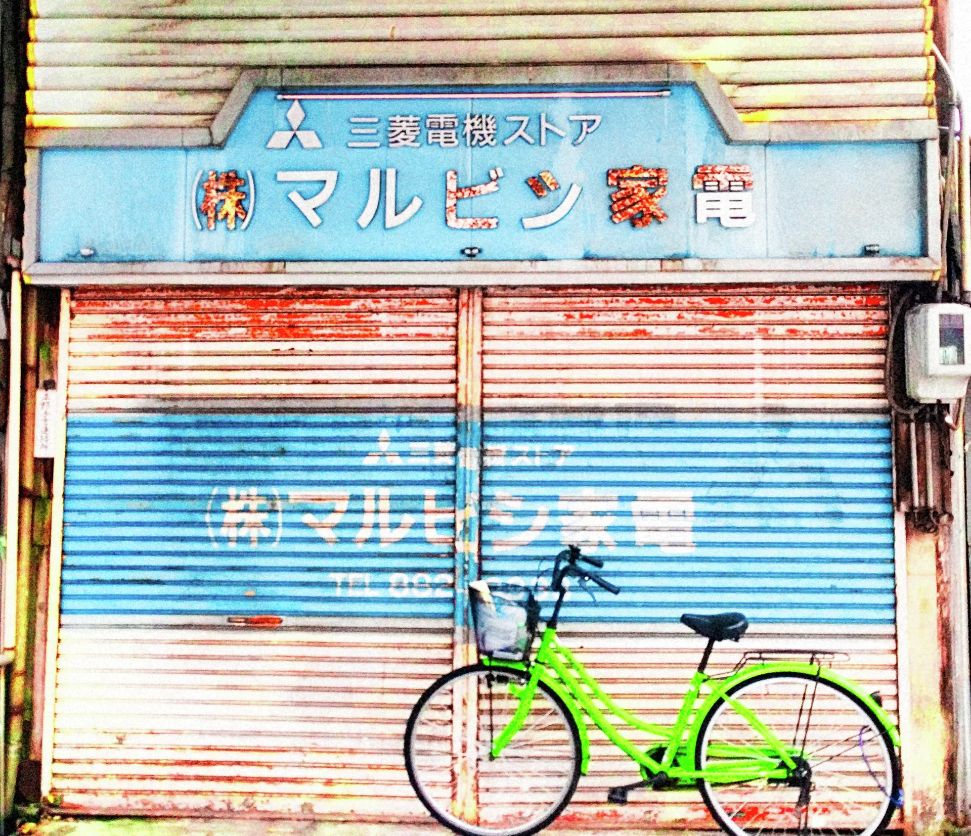 A bright green bicycle leans against a weathered red, white, and blue shutter beneath a pale blue sign.