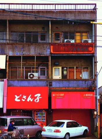 Bright red shop signs sit below weathered upper balconies above two parked cars on a narrow street.