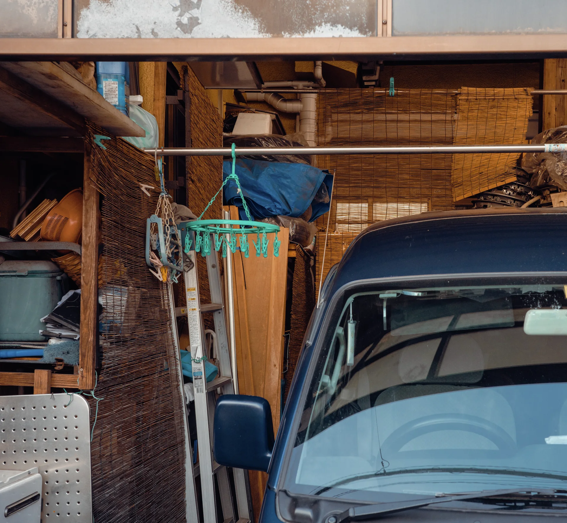 A dark blue van occupies a cluttered garage beside hanging clips, wooden panels and stored materials.