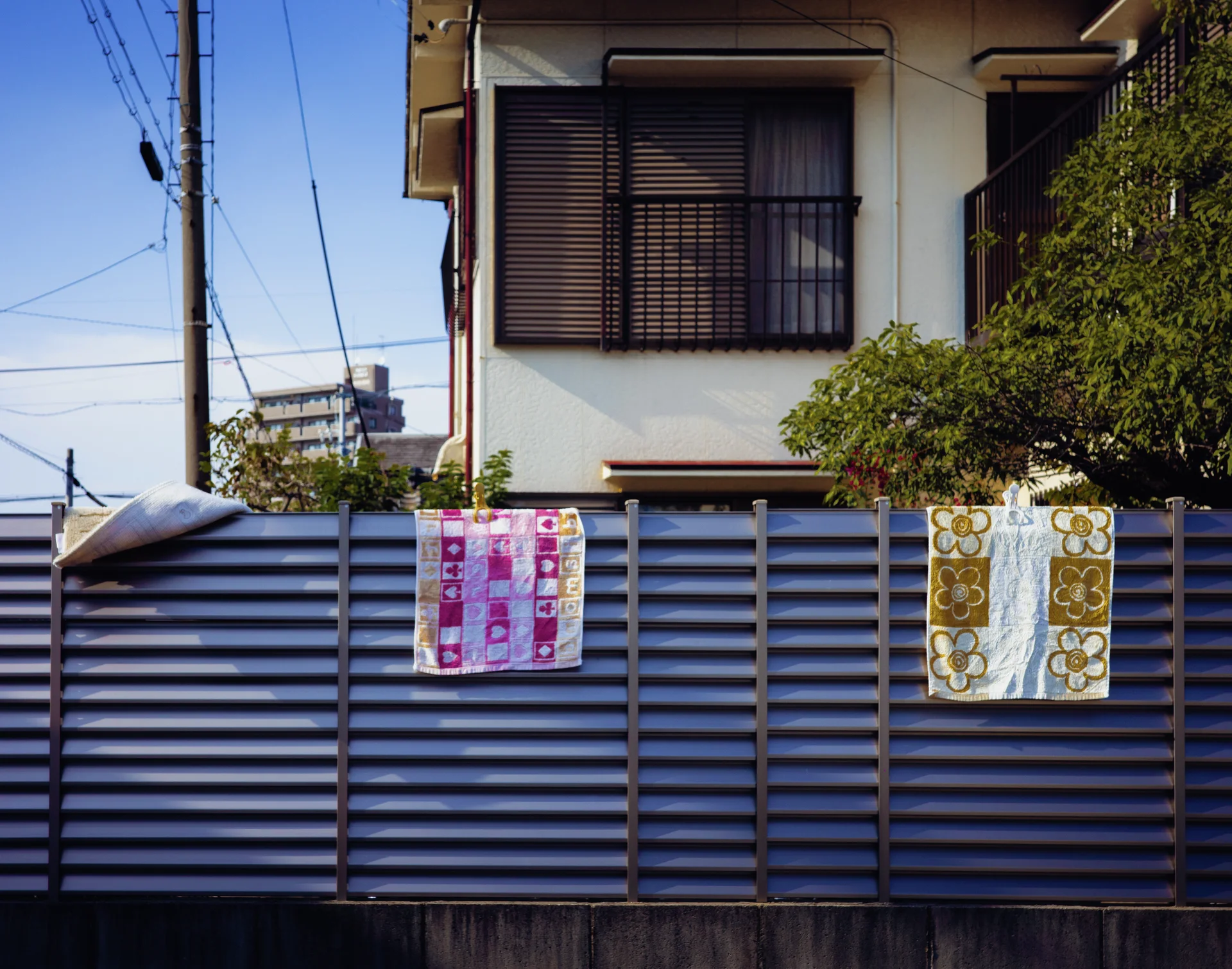 Two patterned cloths hang on a blue slatted fence outside a house with a dark upper window.