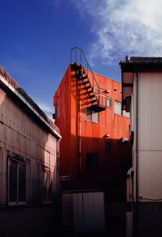 An orange building with a narrow external staircase rises between pale walls beneath blue sky.