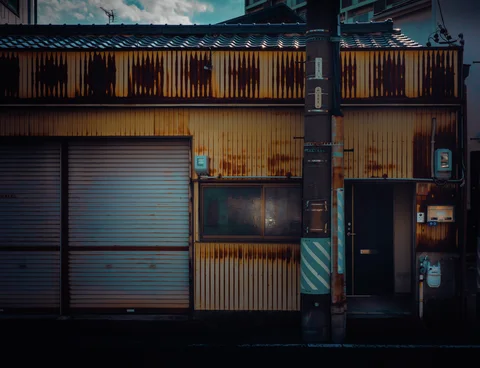 A utility pole stands before a yellow corrugated building with closed shutters, a dark window, and doorway.