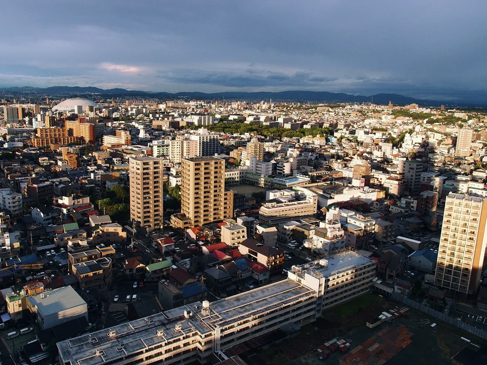 Sunlight illuminates dense city buildings beneath dark gray clouds, with parallel railway tracks in the foreground.