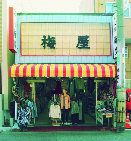 A red and yellow striped awning shades a clothing shop with hanging garments and a figure at its entrance.