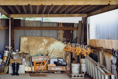 Hanging onions, containers, tools, and rubber boots fill a cluttered shelter beneath corrugated roofing.