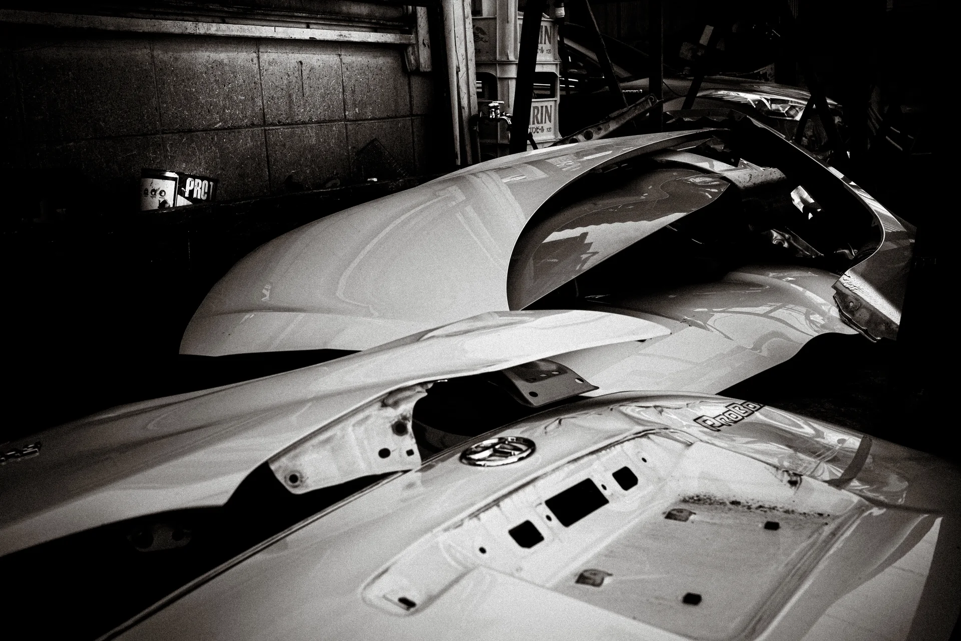 Detached pale vehicle panels lie stacked in a dark workshop in a black and white view.