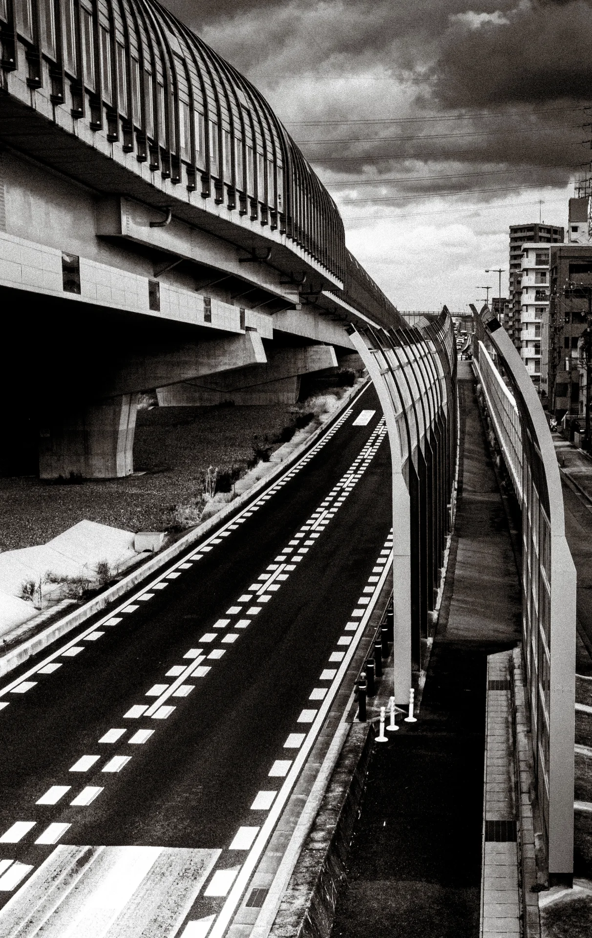 A narrow empty road curves between elevated infrastructure and a tall barrier beneath clouds in black and white.
