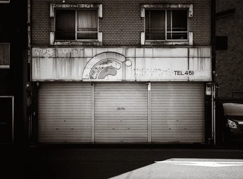Closed metal shutters sit beneath a weathered shop sign and two upstairs windows in black and white.