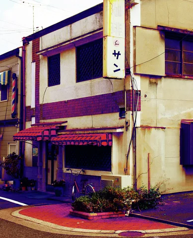 A narrow weathered cream building has red awnings, dark windows, and a vertical sign beside a corner planter.