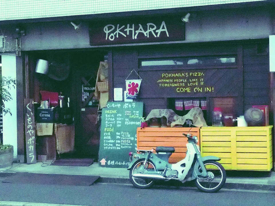A pale turquoise scooter parks outside a dark restaurant with chalkboard menus and yellow wooden barriers.