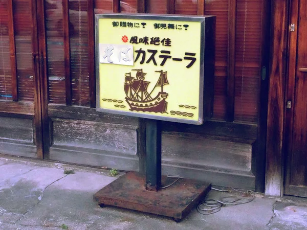 A cream freestanding sign with Japanese lettering and a sailing ship illustration stands beside dark wooden windows.