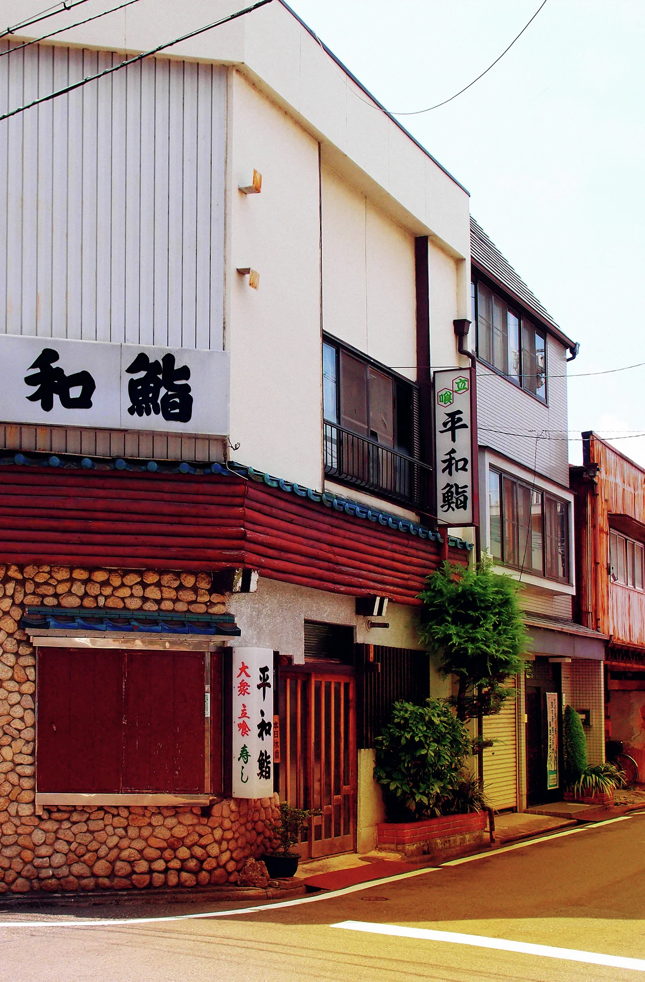 A narrow corner restaurant has pale upper siding, a red awning and stone-patterned lower walls.