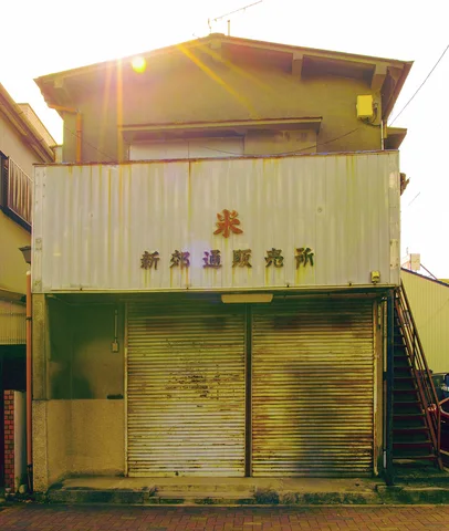 Sunlight glints above a corrugated building with faded Japanese lettering and two closed metal shutters.
