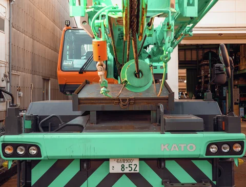 A green mobile crane has an orange cab, hanging hooks, and bold diagonal stripes across its front.