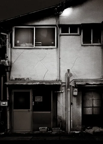 Two narrow entrances sit beneath mismatched upper windows on a weathered two-story facade in black and white.