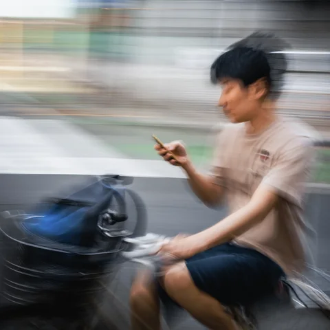 A cyclist holds a smartphone beside a loaded front basket against a horizontally blurred street.