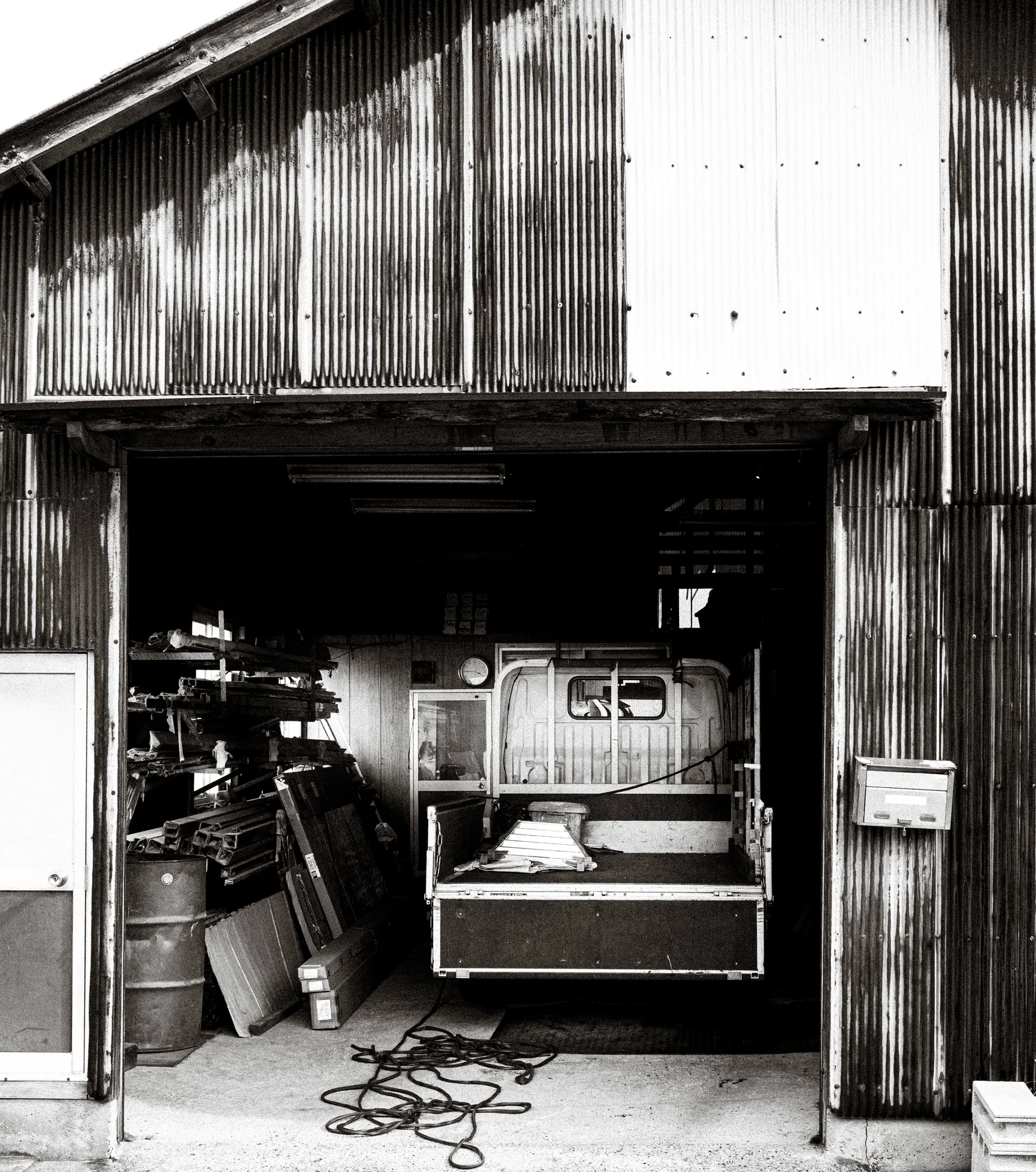 An open corrugated workshop reveals stacked materials and machinery with a coiled cable outside in black and white.