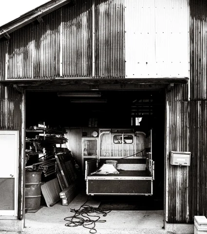 An open corrugated workshop reveals stacked materials and machinery with a coiled cable outside in black and white.