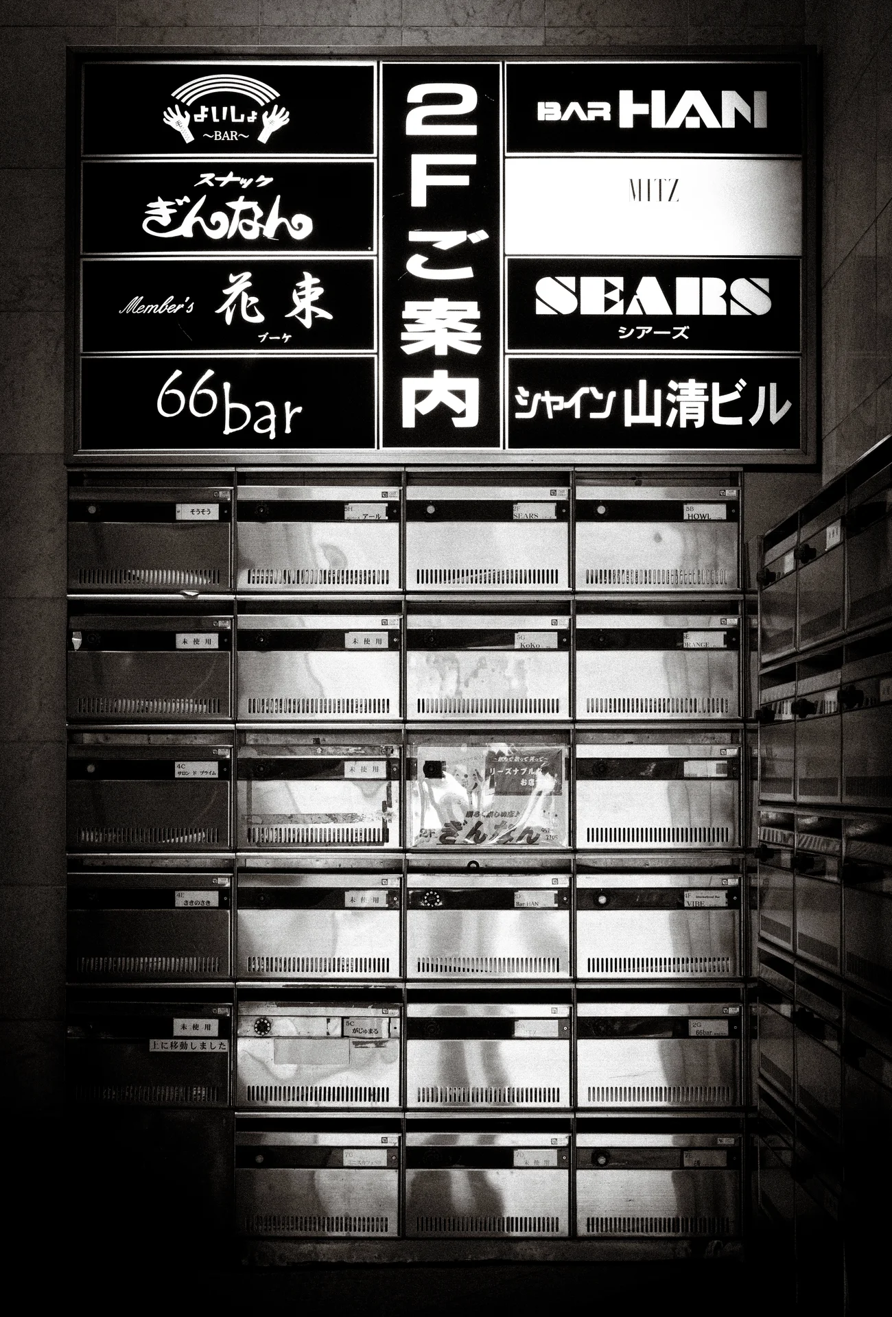 A tall building directory of dark signs and reflective nameplates fills a shadowed monochrome entrance.