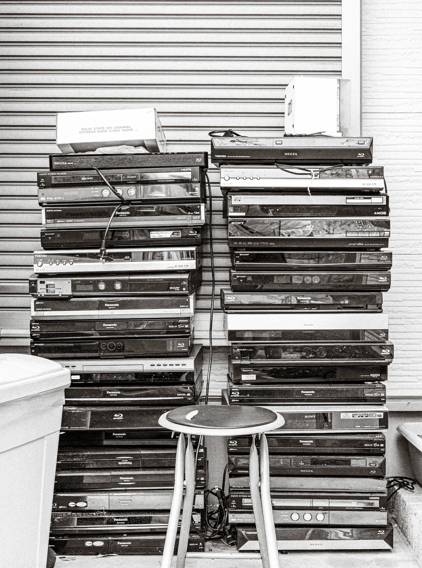 Stacks of shallow wooden trays stand against closed shutters behind a small round stool in black and white.