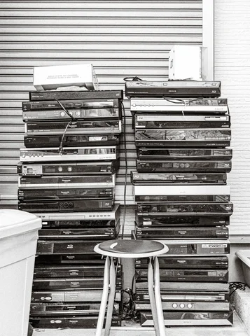 Stacks of shallow wooden trays stand against closed shutters behind a small round stool in black and white.