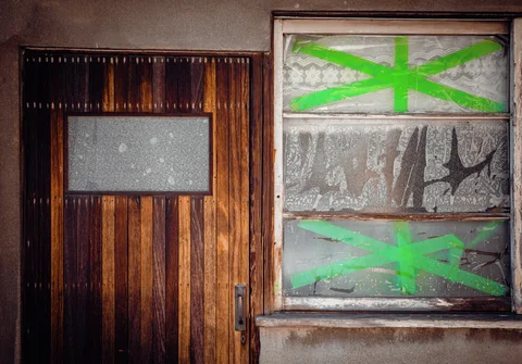 Bright green tape crosses damaged window panes beside a wooden door with a small frosted panel.