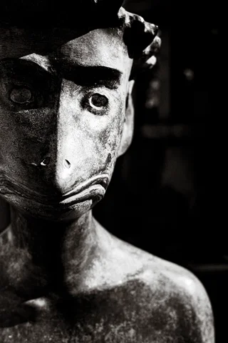 A weathered sculpted face with round eyes emerges from deep shadow in black and white.
