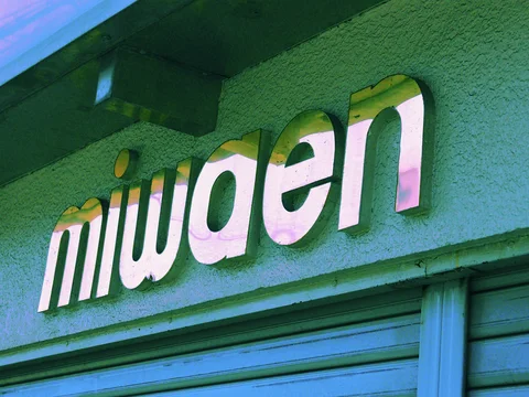 Raised lowercase miwaen letters cast shadows across a textured green wall above a closed blue shutter.