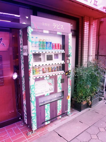 A tall drink vending machine stands beside a purple-lit entrance and a leafy potted shrub.