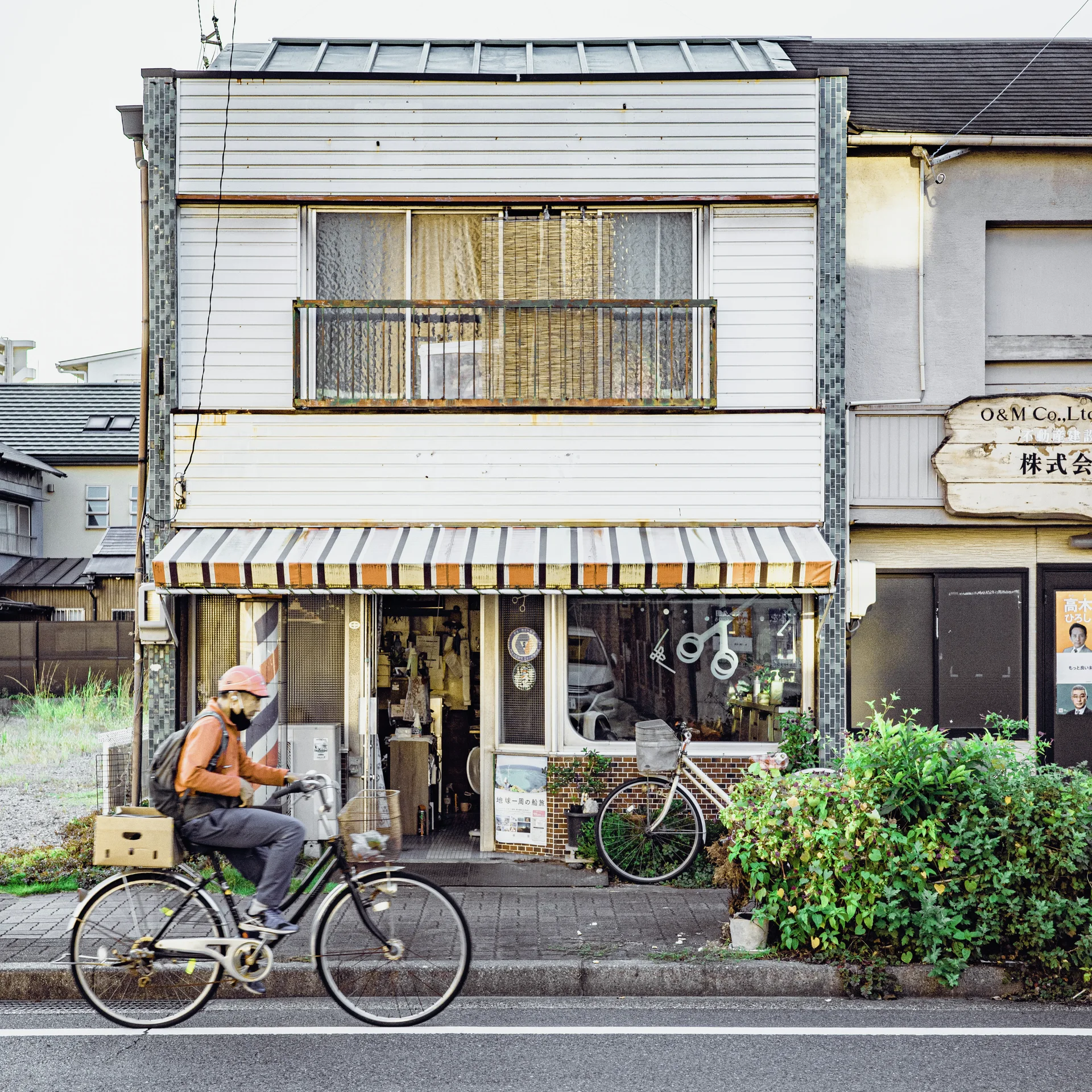 A cyclist rides past a narrow white storefront with a striped awning and leafy plants.