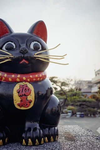 A black cat figurine with long gold whiskers and a red collar sits on a low wall.