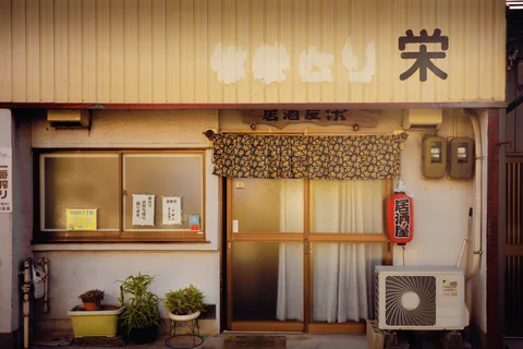 A small pale storefront has curtained doors, a patterned valance, potted plants, and an outdoor air conditioner.