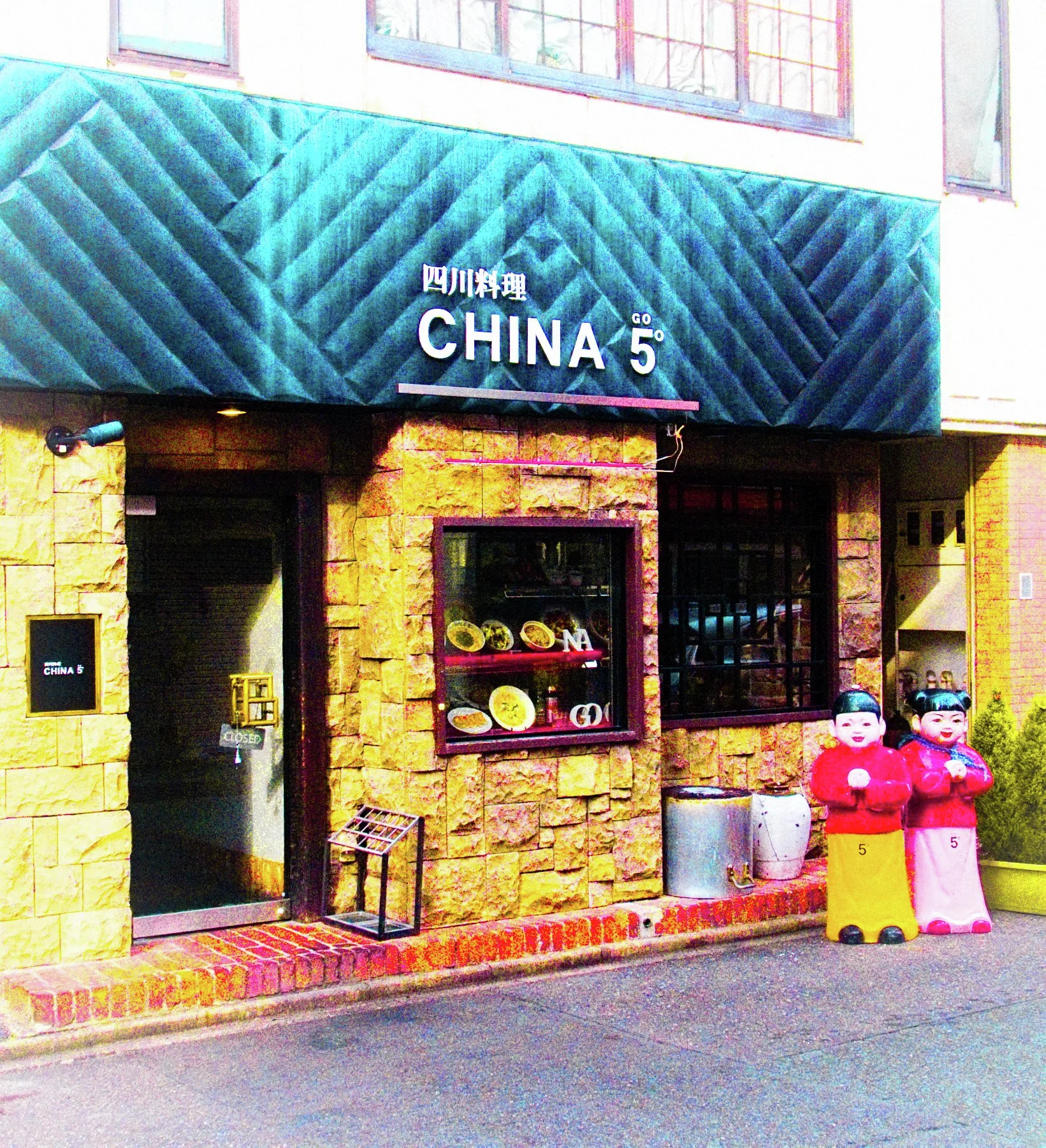 A restaurant entrance has yellow stone walls, a dark turquoise awning, and two bright cartoon figures.